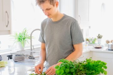Person preparing a sandwich in a bright kitchen, adding sliced vegetables to the bread with fresh herbs on the counter.