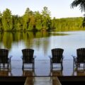 A view looking out on a lake at sunset with a wooden floating dock and four lounge chairs looking out.