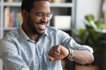 A man wearing glasses and a patterned button-up shirt sits in front of a laptop. He smiles and looks at his wristwatch.
