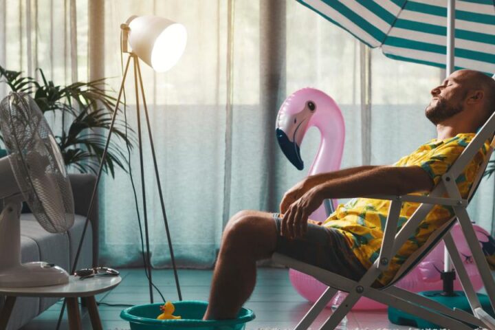 Man relaxing indoors on a lounge chair under a striped umbrella, cooling off with a fan, creating a playful home.