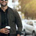 Man wearing sunglasses walks on city street holding coffee cup, with parked cars and buildings in soft sunlight.