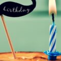 One lit candle is on a small, round, unfrosted cake. Next to it, a mustache-shaped cake topper says, "Happy Birthday."