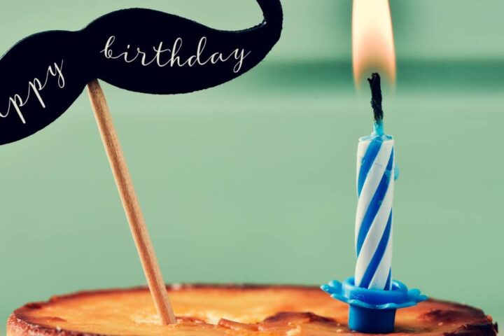 One lit candle is on a small, round, unfrosted cake. Next to it, a mustache-shaped cake topper says, "Happy Birthday."