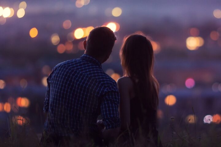 A couple sitting closely on a hill at dusk, overlooking a city with glowing lights, sharing a romantic moment together.
