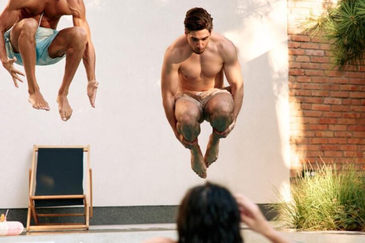 Two young men jumping midair into a backyard swimming pool while friends watch and relax nearby.