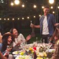 Group of friends enjoying an outdoor dinner party at night, eating, and socializing under warm string lights in a backyard setting.