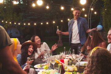Group of friends enjoying an outdoor dinner party at night, eating, and socializing under warm string lights in a backyard setting.