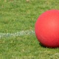 A red kickball resting on green grass field near a white boundary line, suggesting an outdoor recreational play.