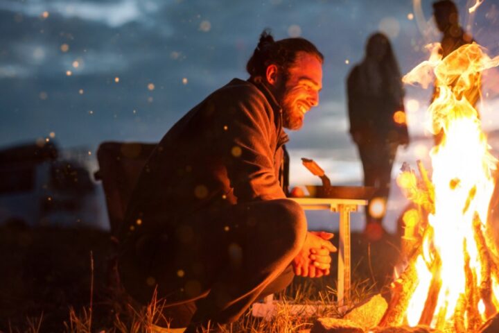 A smiling bearded man sitting beside a bright bonfire at night, with orange flames lighting his face.