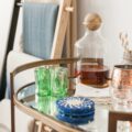 A round glass bar cart holds a corked decanter, green shot glasses, blue coasters, striped straws, and copper mules.