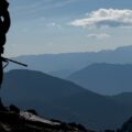 The silhouette of a man with a rifle standing on a ridge during the day, with a blue sky and mountain ridges behind him.