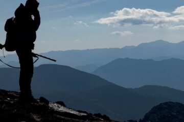 The silhouette of a man with a rifle standing on a ridge during the day, with a blue sky and mountain ridges behind him.