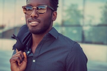 A man wearing sunglasses and a patterned button-up shirt holds a jacket over one shoulder outside a glass building.