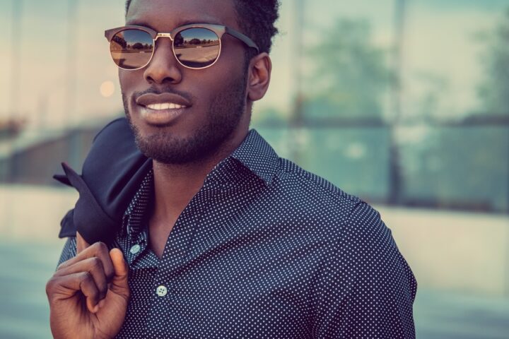 A man wearing sunglasses and a patterned button-up shirt holds a jacket over one shoulder outside a glass building.