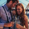 A man and woman standing close by the water while holding glasses under string lights with a bridge behind them.