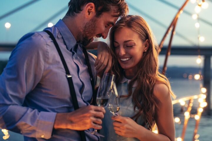 A man and woman standing close by the water while holding glasses under string lights with a bridge behind them.