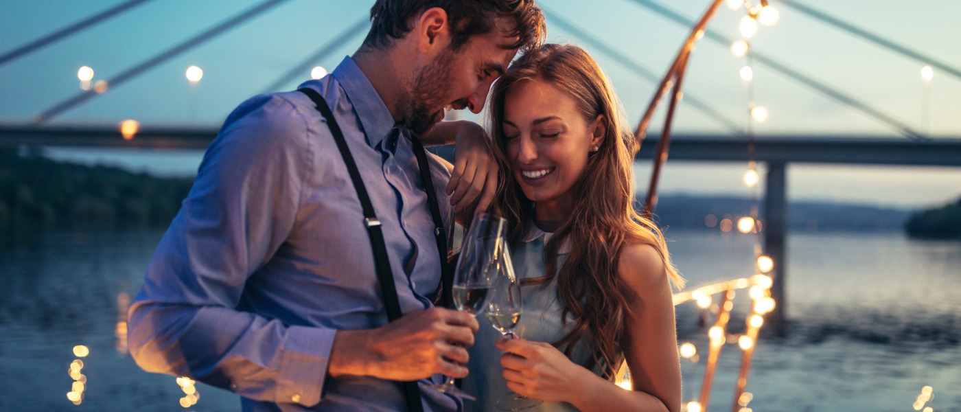 A man and woman standing close by the water while holding glasses under string lights with a bridge behind them.