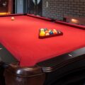 A luxurious home billiards room featuring a black pool table with bright red felt, racked billiard balls, and modern accent lighting along a dark brick wall.
