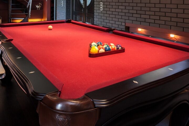 A luxurious home billiards room featuring a black pool table with bright red felt, racked billiard balls, and modern accent lighting along a dark brick wall.