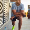 A young man in workout clothes stretching in an urban area. He has his water bottle on the floor next to him.