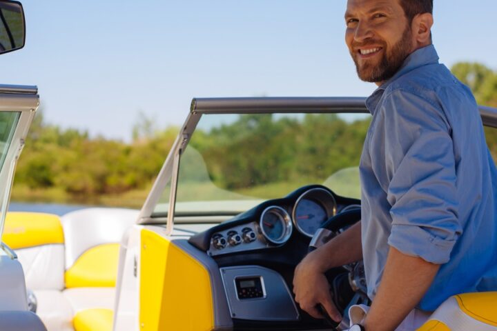 A man with a beard and a blue shirt, piloting a speedboat and looking back over his shoulder and smiling.