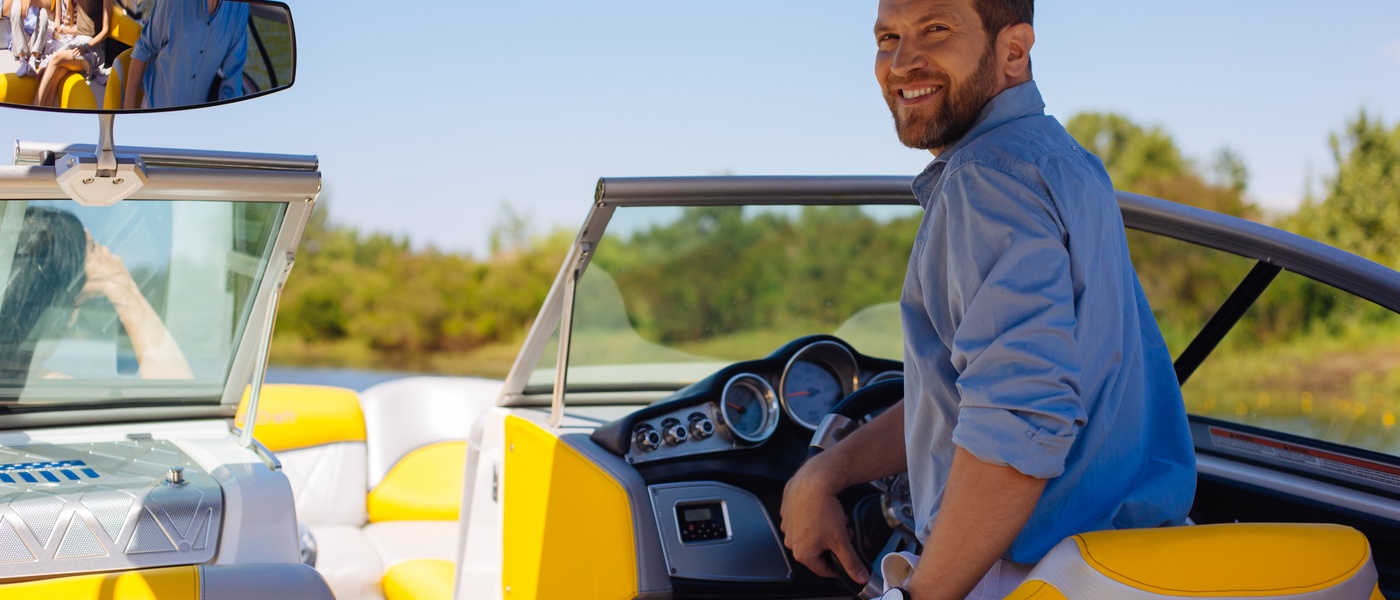 A man with a beard and a blue shirt, piloting a speedboat and looking back over his shoulder and smiling.