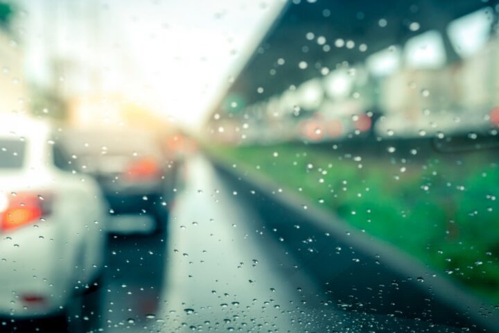 A view of raindrops on a car window. The car appears parked at a train station with other cars nearby.