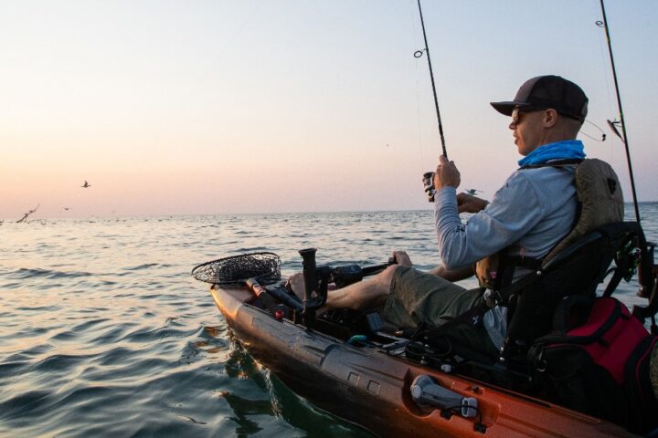 A man fishing from a kayak on calm open water at sunrise, with fishing rods and gear on the boat and birds visible nearby.