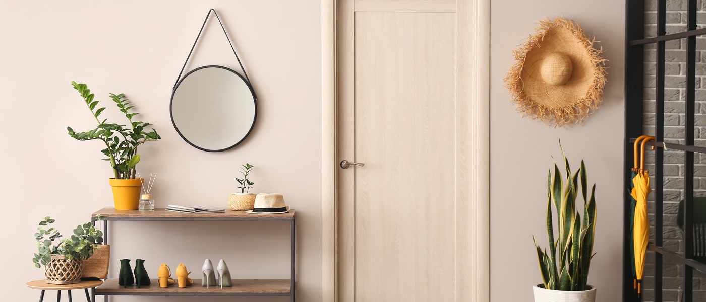 A narrow console table with a shelf holds shoes and plants. Above it hangs a round mirror; beside it is a door.