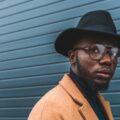 A stylish man in a tan overcoat, black hat, and glasses stands in front of a blue metal wall while looking away.