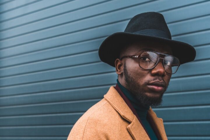 A stylish man in a tan overcoat, black hat, and glasses stands in front of a blue metal wall while looking away.