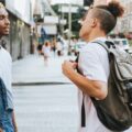 Two men standing at a city crosswalk on a busy street, one wearing denim overalls and the other carrying a backpack.