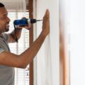 A man using a cordless power drill to make a hole in an interior house wall while holding the surface steady.