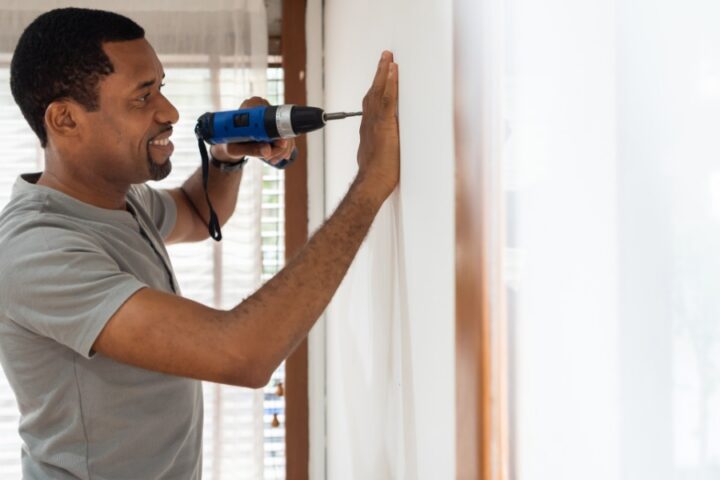 A man using a cordless power drill to make a hole in an interior house wall while holding the surface steady.