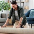 A smiling man kneeling on a living room floor while unrolling a beige rug with a sofa and blue chair behind him.