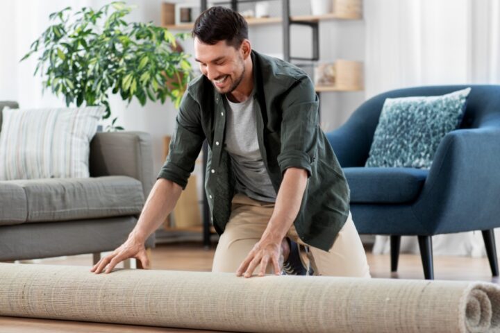 A smiling man kneeling on a living room floor while unrolling a beige rug with a sofa and blue chair behind him.