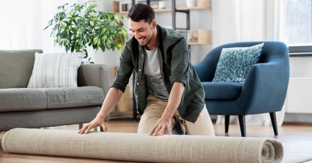 A smiling man kneeling on a living room floor while unrolling a beige rug with a sofa and blue chair behind him.