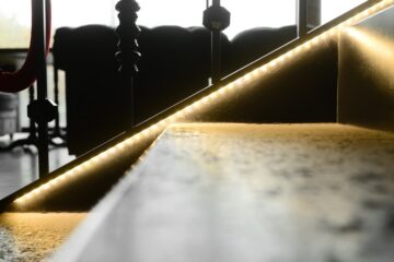A glowing LED strip traces a dark metal staircase as sunlight enters through the windows in the background.