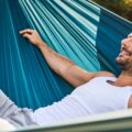 A man in a white tank top and gray shorts lying in a blue hammock on a sunny lawn with shrubs behind him.
