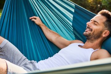 A man in a white tank top and gray shorts lying in a blue hammock on a sunny lawn with shrubs behind him.