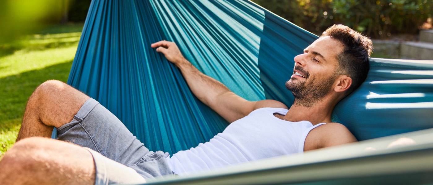 A man in a white tank top and gray shorts lying in a blue hammock on a sunny lawn with shrubs behind him.