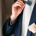 A white flower boutonniere pinned to the lapel of a dark suit jacket with a bow tie and vest visible.