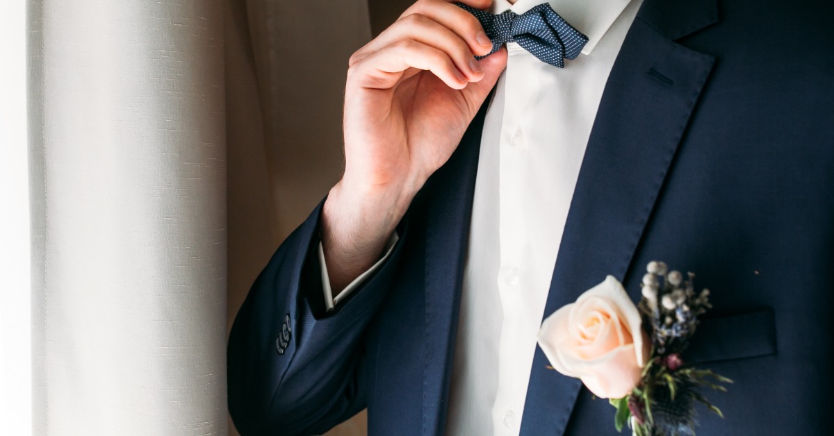A white flower boutonniere pinned to the lapel of a dark suit jacket with a bow tie and vest visible.