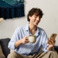 A man in a blue shirt sitting on a sofa, holding a mug and looking at a phone near plants and wall art.