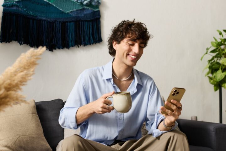 A man in a blue shirt sitting on a sofa, holding a mug and looking at a phone near plants and wall art.