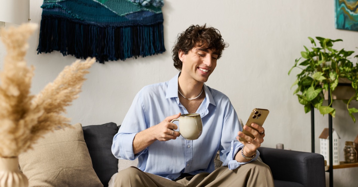 A man in a blue shirt sitting on a sofa, holding a mug and looking at a phone near plants and wall art.