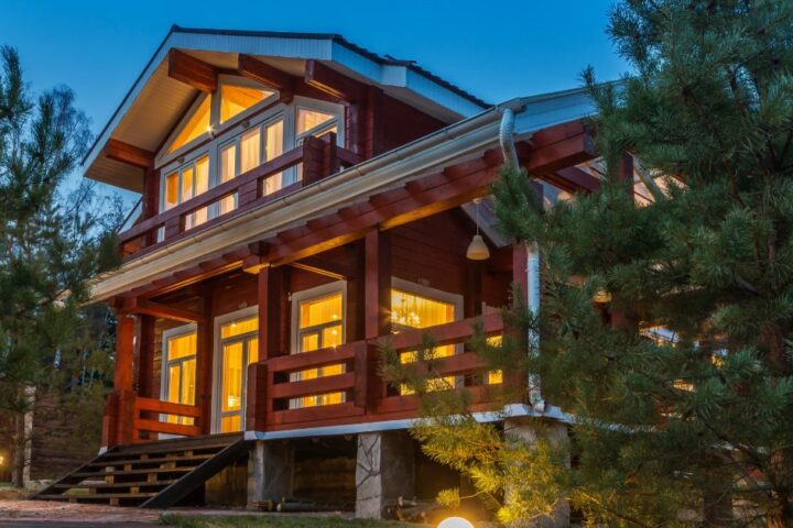 A two-story wooden cabin glows with warm interior lights as tall pine trees surround it beneath a blue evening sky.