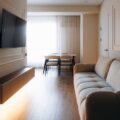 Modern narrow living room with a beige sofa, wall-mounted television, floating cabinet, and dining area by a window.